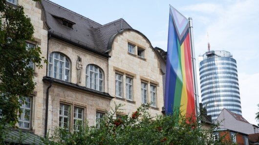 Hauptgebäude der Friedrich-Schiller-Universität Jena mit Regenbogenfahne und Jentower im Hintergrund